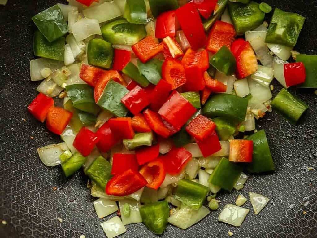 Chopped red and green bell peppers and onions are being sautéed in a pan. The vegetables are vibrant and colorful, and the pan has a textured non-stick surface.